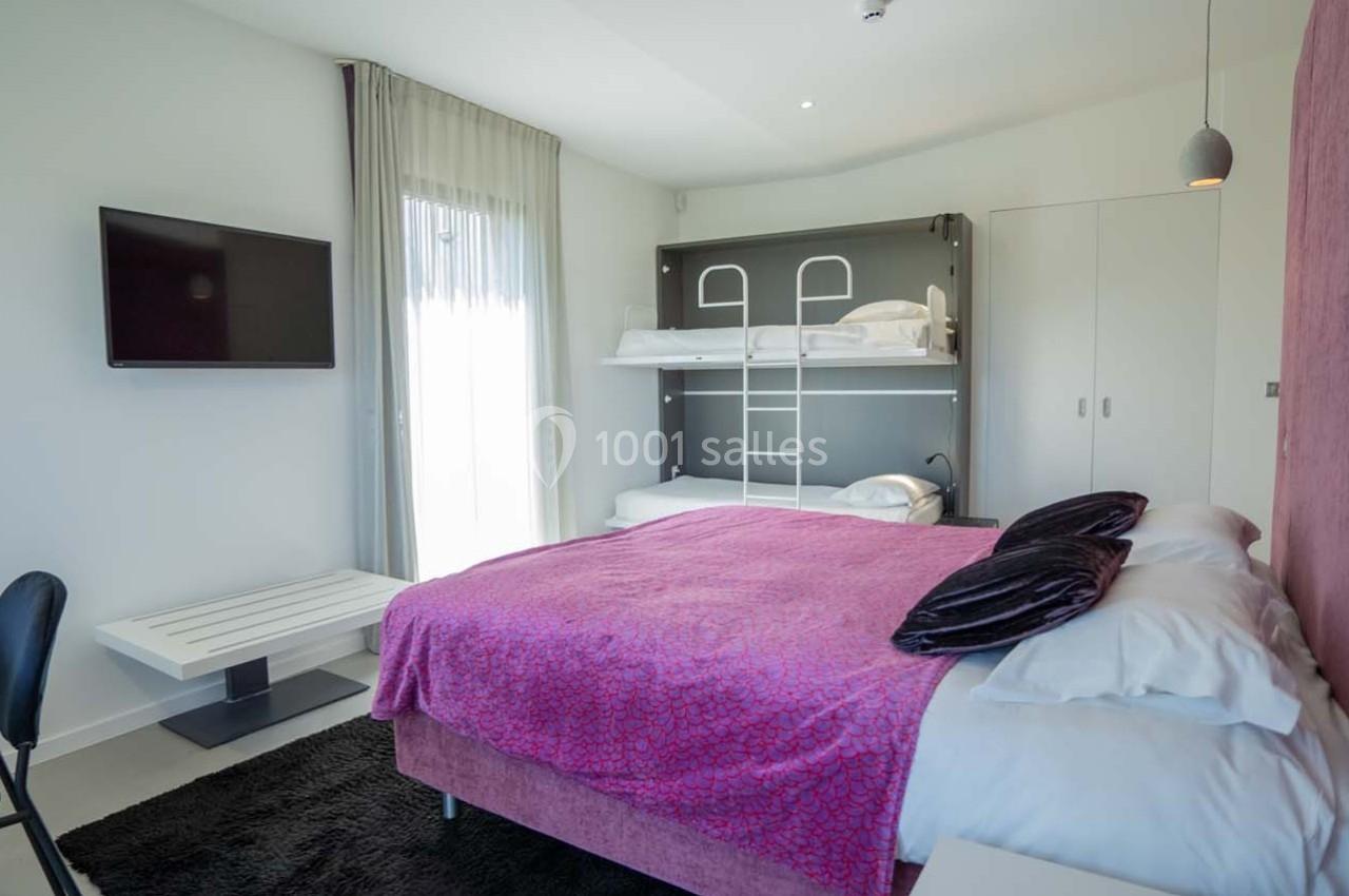 Chambre moderne avec lit double, lits superposés, télévision murale, rideaux translucides et mobilier minimaliste.