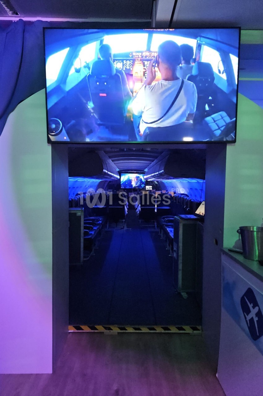 Vue intérieure d'un simulateur d'avion avec des sièges alignés et un écran montrant un cockpit en fonctionnement.
