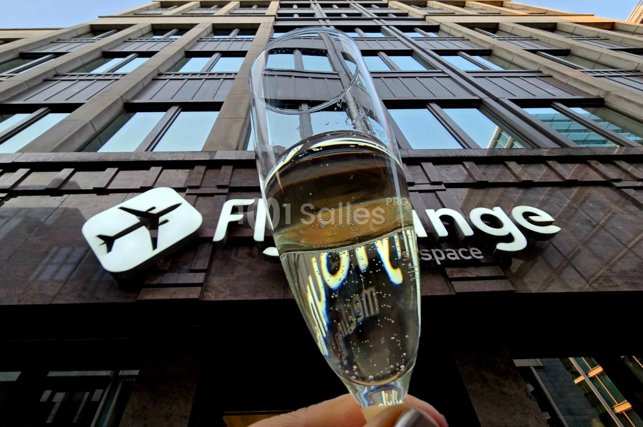 Verre de champagne tenu devant la façade d'un bâtiment avec une enseigne ’Fly’ et un logo d'avion.