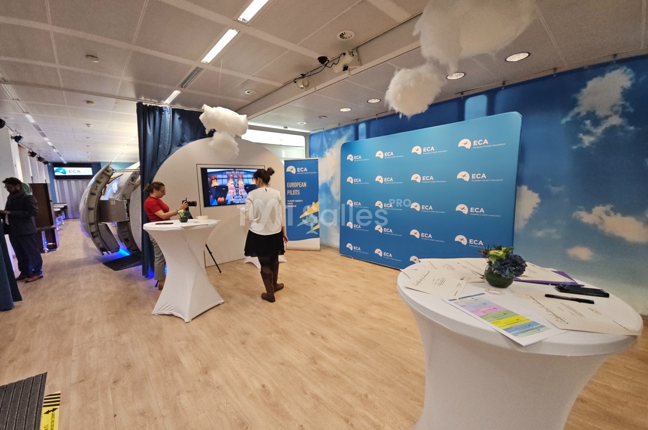Salle d'exposition avec des stands, des écrans interactifs et des tables hautes, décorée de nuages suspendus.