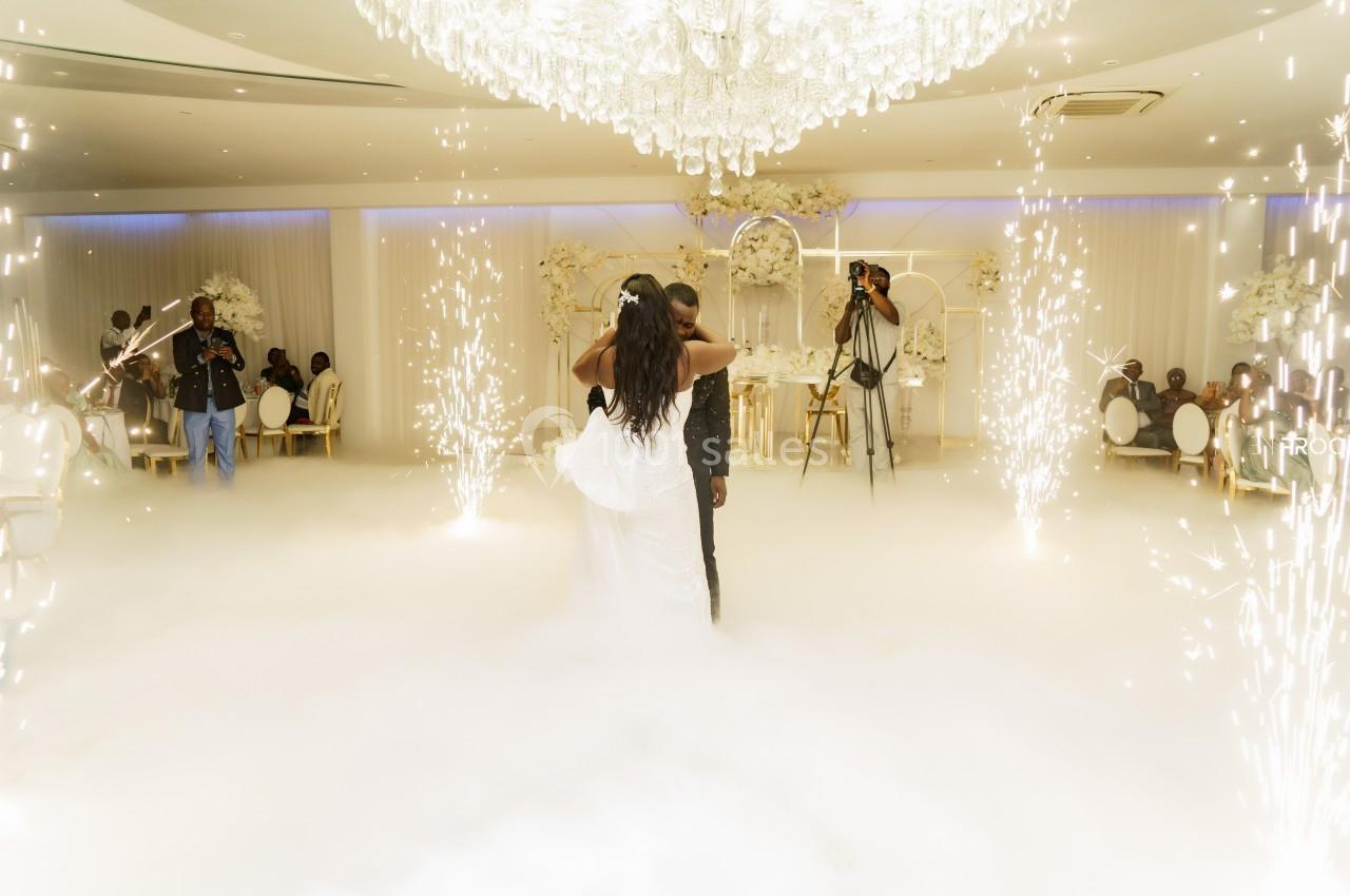 Un couple danse sous un lustre imposant, entouré de fumée et d'étincelles, dans une salle décorée en blanc.