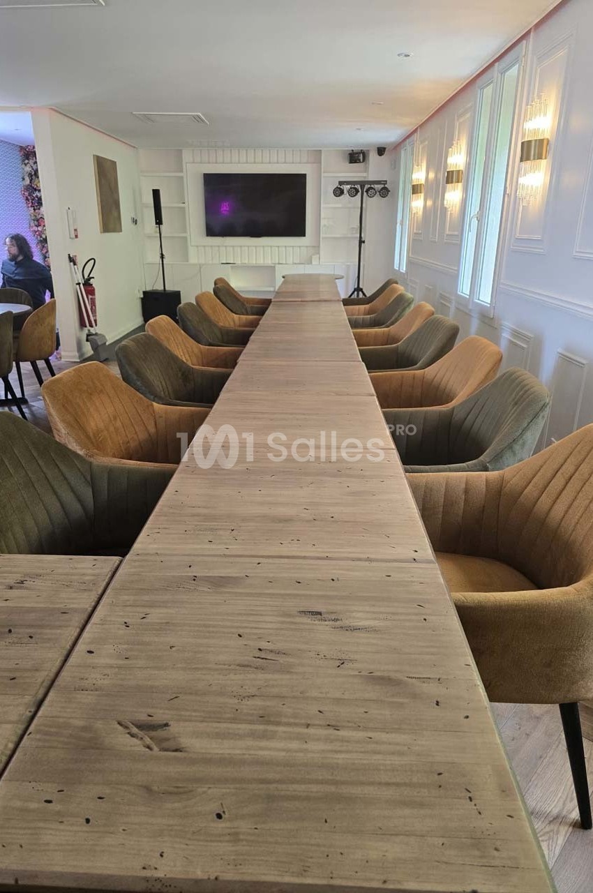 Grande table en bois entourée de fauteuils rembourrés dans une salle lumineuse avec écran et éclairages en arrière-plan.
