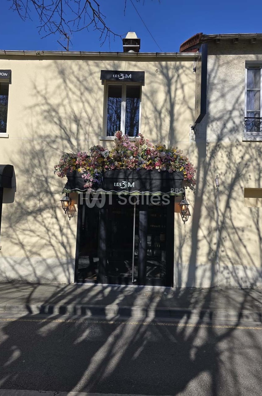 Façade d'un bâtiment avec une porte noire, une enseigne ’Les 3M’ et des fleurs colorées ornant le haut.