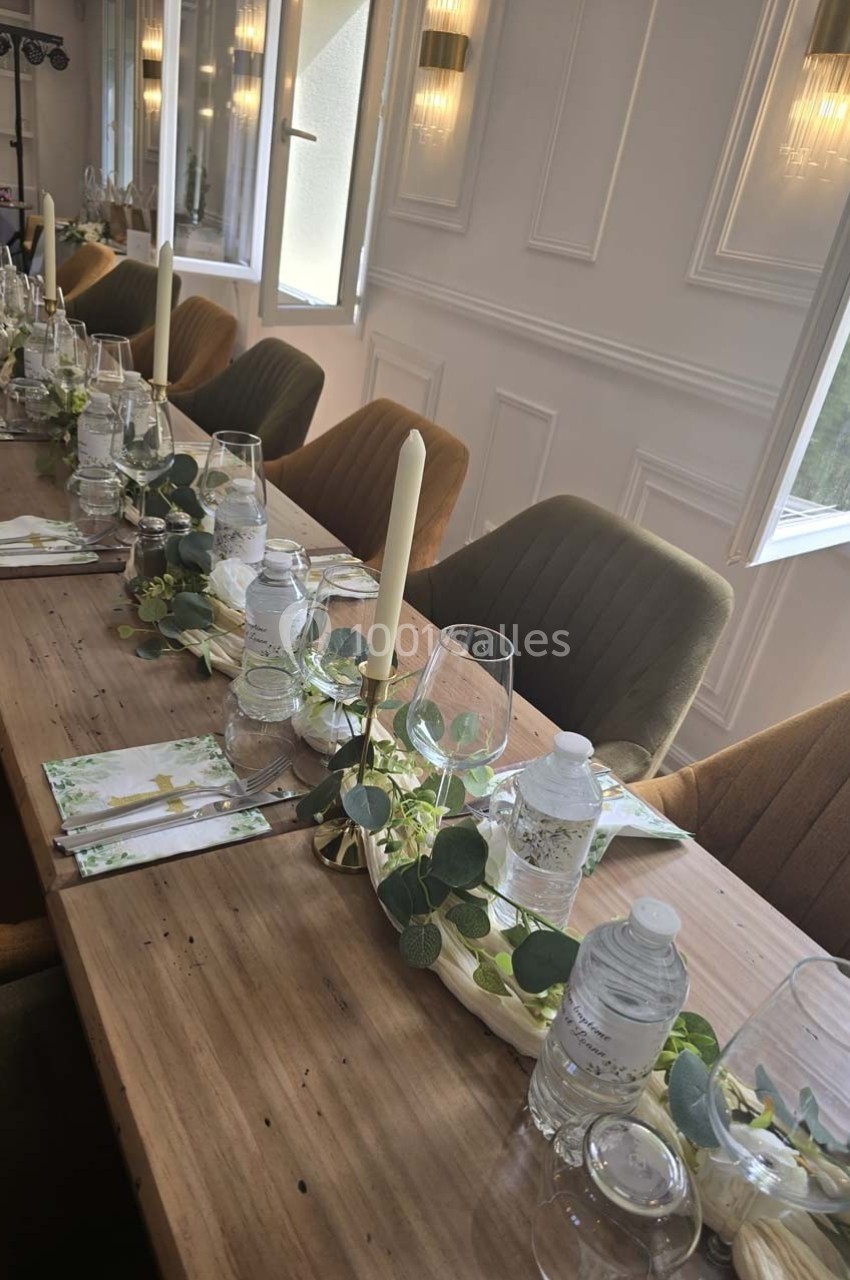 Table en bois décorée avec des bougies, feuillage, serviettes pliées et bouteilles d'eau, dans une salle lumineuse.