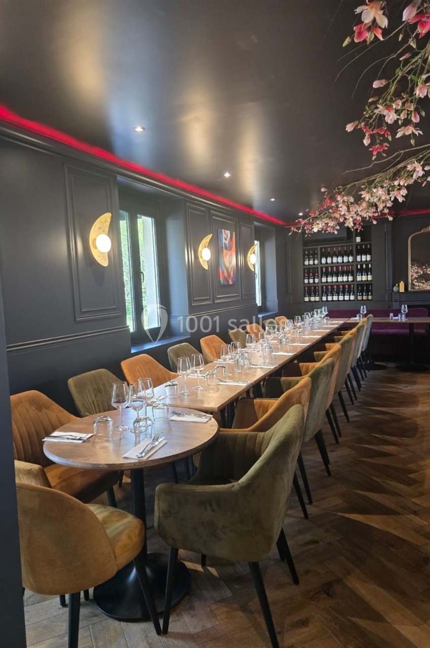 Salle de restaurant élégante avec une longue table dressée, chaises en velours et décoration florale au plafond.
