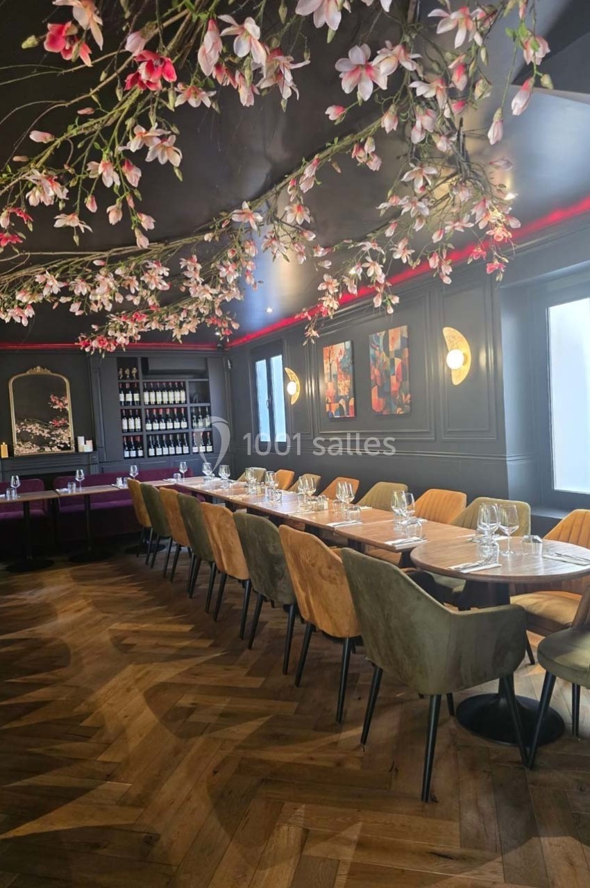 Salle de restaurant élégante avec une longue table en bois, chaises colorées et décoration florale suspendue.