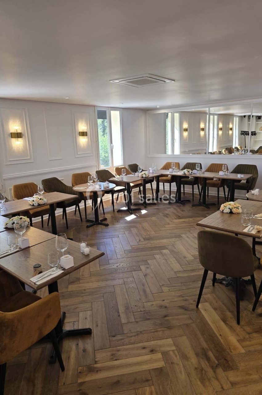 Salle lumineuse avec tables en bois dressées, chaises rembourrées et parquet en chevrons, prête pour un repas ou une réunion.