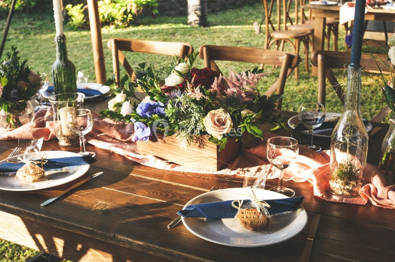 Table en bois dressée pour un repas en extérieur, décorée de fleurs, bougies et serviettes bleues.