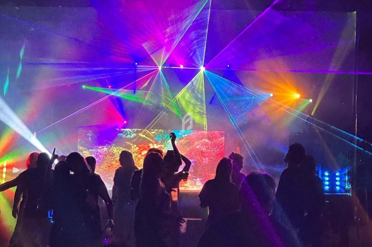Personnes dansant dans une salle sombre illuminée par des faisceaux lumineux colorés et des effets visuels.