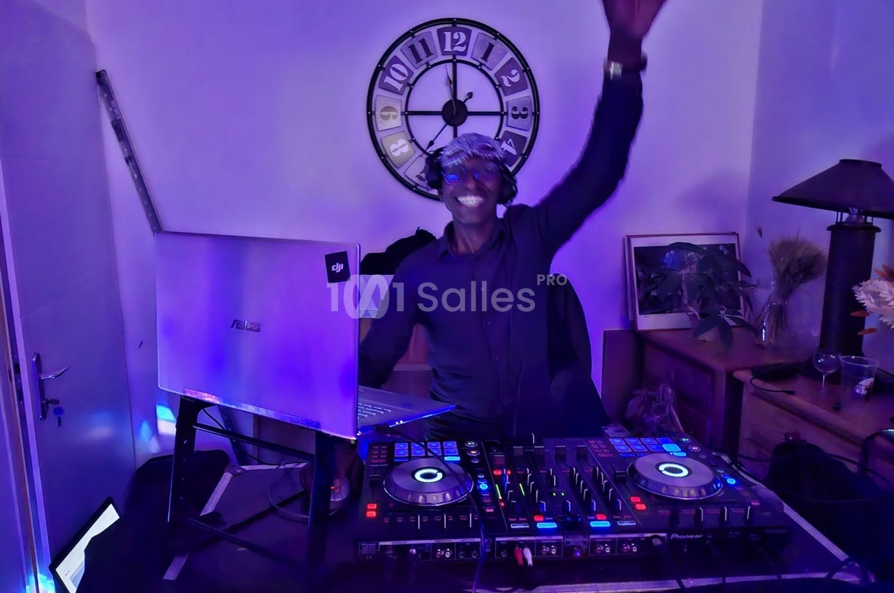 Un DJ souriant mixe de la musique sur une table de mixage dans une pièce éclairée par des lumières colorées.