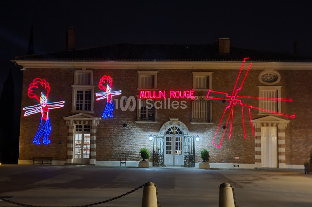 Façade en briques éclairée la nuit avec des néons rouges représentant des danseuses et un moulin, inscription ’Moulin Rouge’.