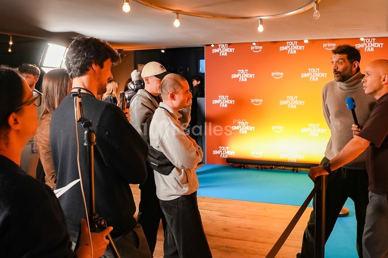 Des personnes discutent devant un fond orange avec un logo, dans un espace éclairé avec un tapis bleu.