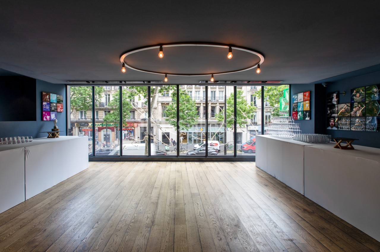 Salle lumineuse avec parquet, grandes baies vitrées donnant sur une rue urbaine, et comptoirs blancs alignés.