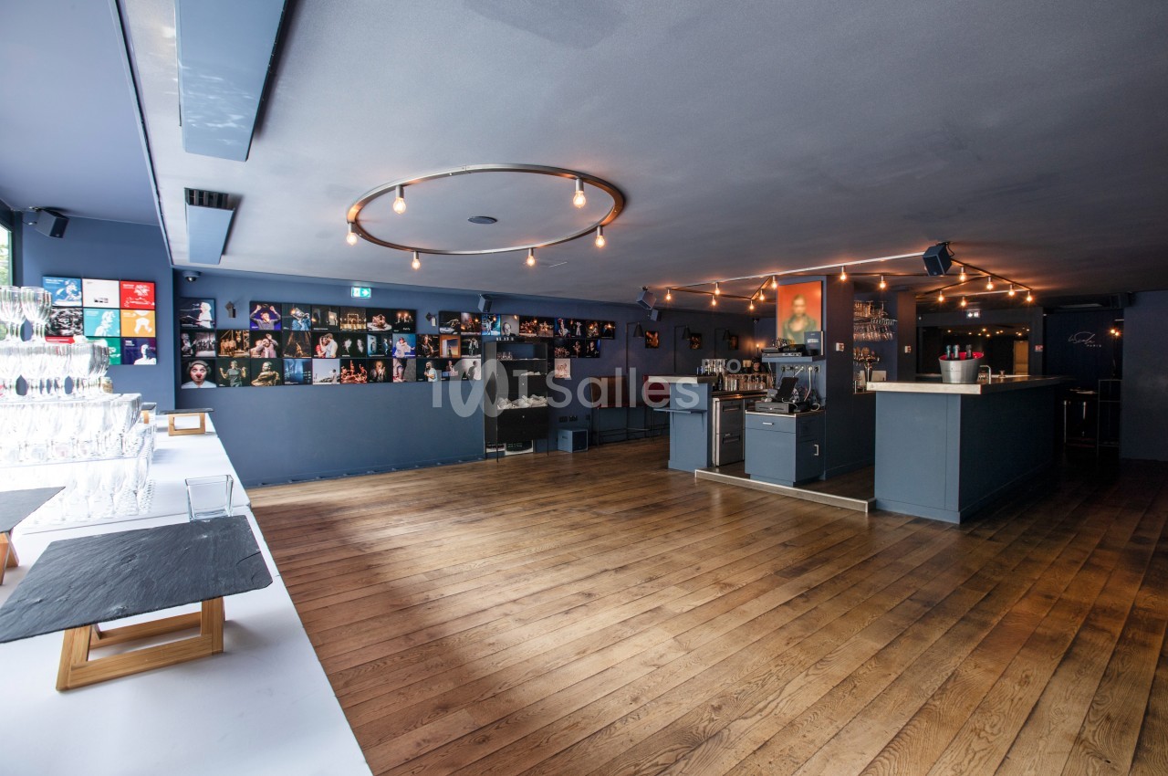 Salle moderne avec parquet en bois, bar à droite, étagères décorées au fond et table avec verres à gauche.