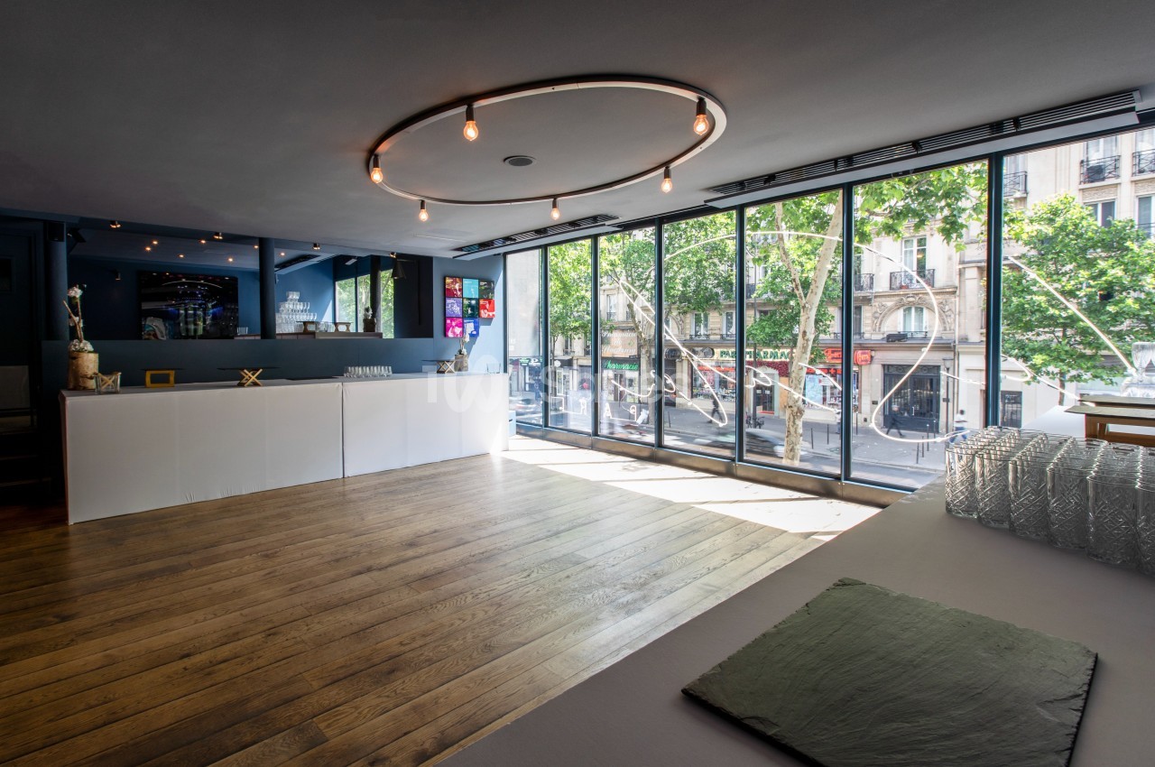 Salle lumineuse avec parquet en bois, grande baie vitrée donnant sur une rue urbaine et comptoir blanc moderne.