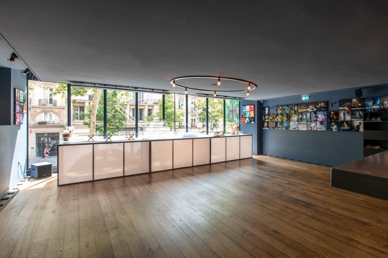 Salle lumineuse avec parquet en bois, grandes baies vitrées donnant sur la rue et étagères décorées sur le mur.