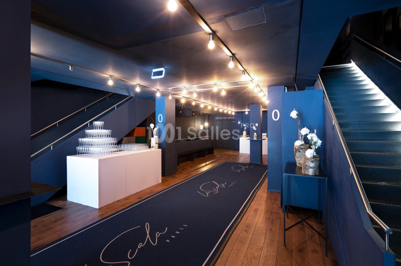Hall d'entrée moderne avec éclairage suspendu, tapis bleu marqué ’La Scala’, escalier et tables décorées.
