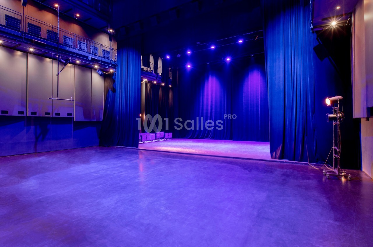 Salle de spectacle vide avec scène éclairée par des projecteurs violets, entourée de rideaux noirs.