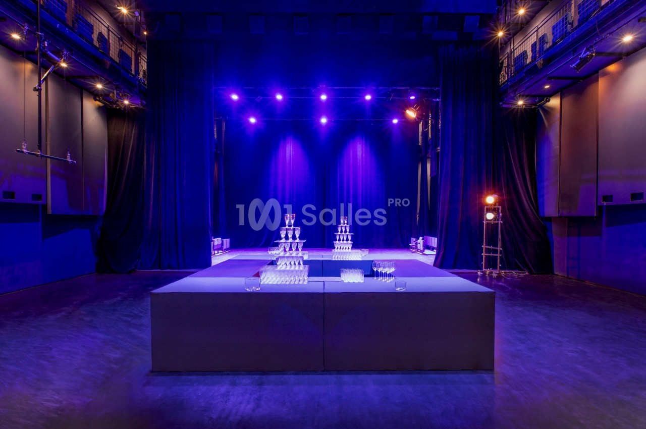 Salle de spectacle avec éclairage violet, scène en arrière-plan et tables décorées de pyramides de verres au premier plan.