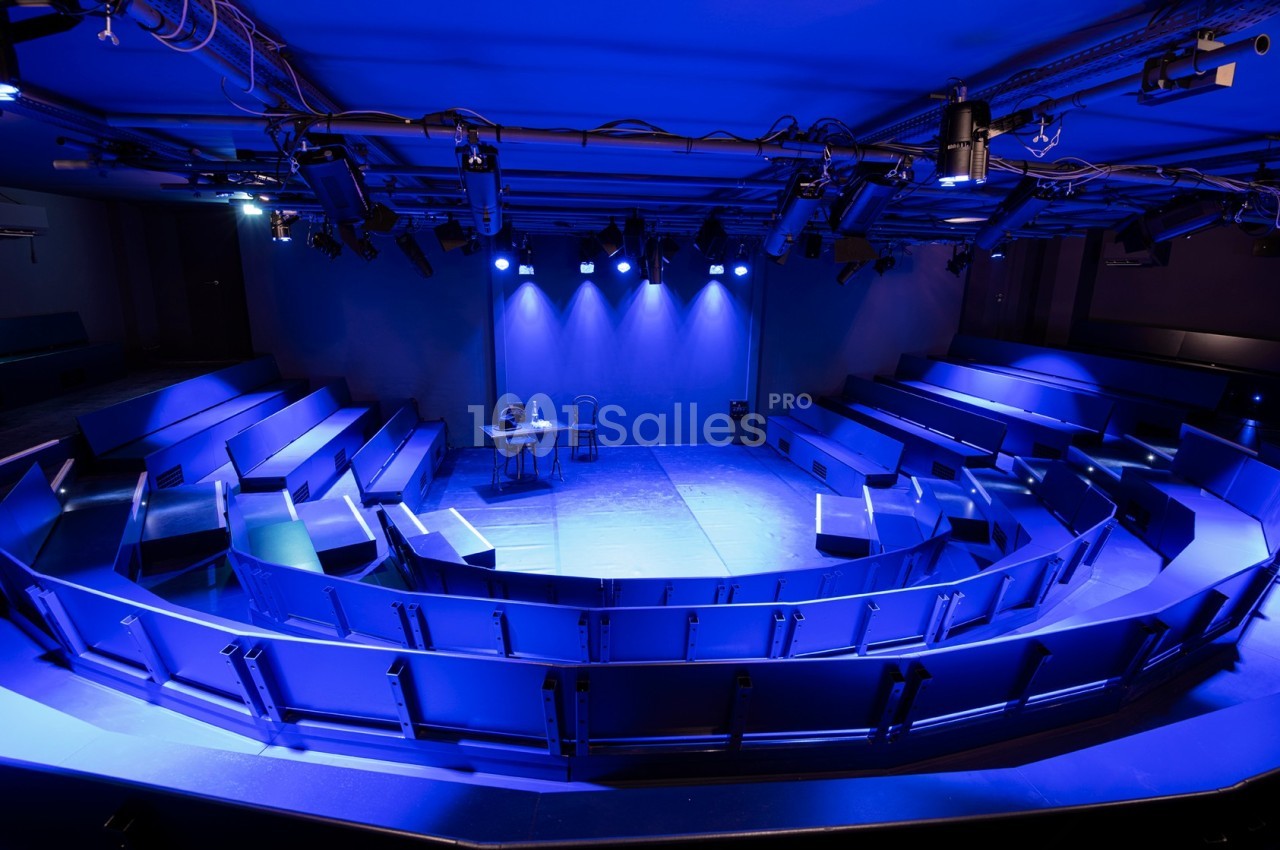Salle de spectacle vide avec gradins en arc de cercle, éclairage bleu et une table avec deux chaises sur scène.