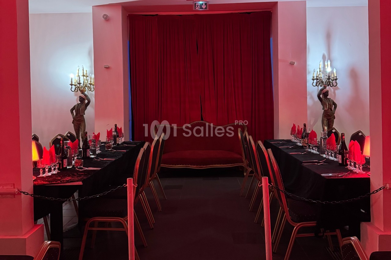 Salle décorée en rouge avec deux longues tables dressées, un canapé central et des statues encadrant des rideaux.