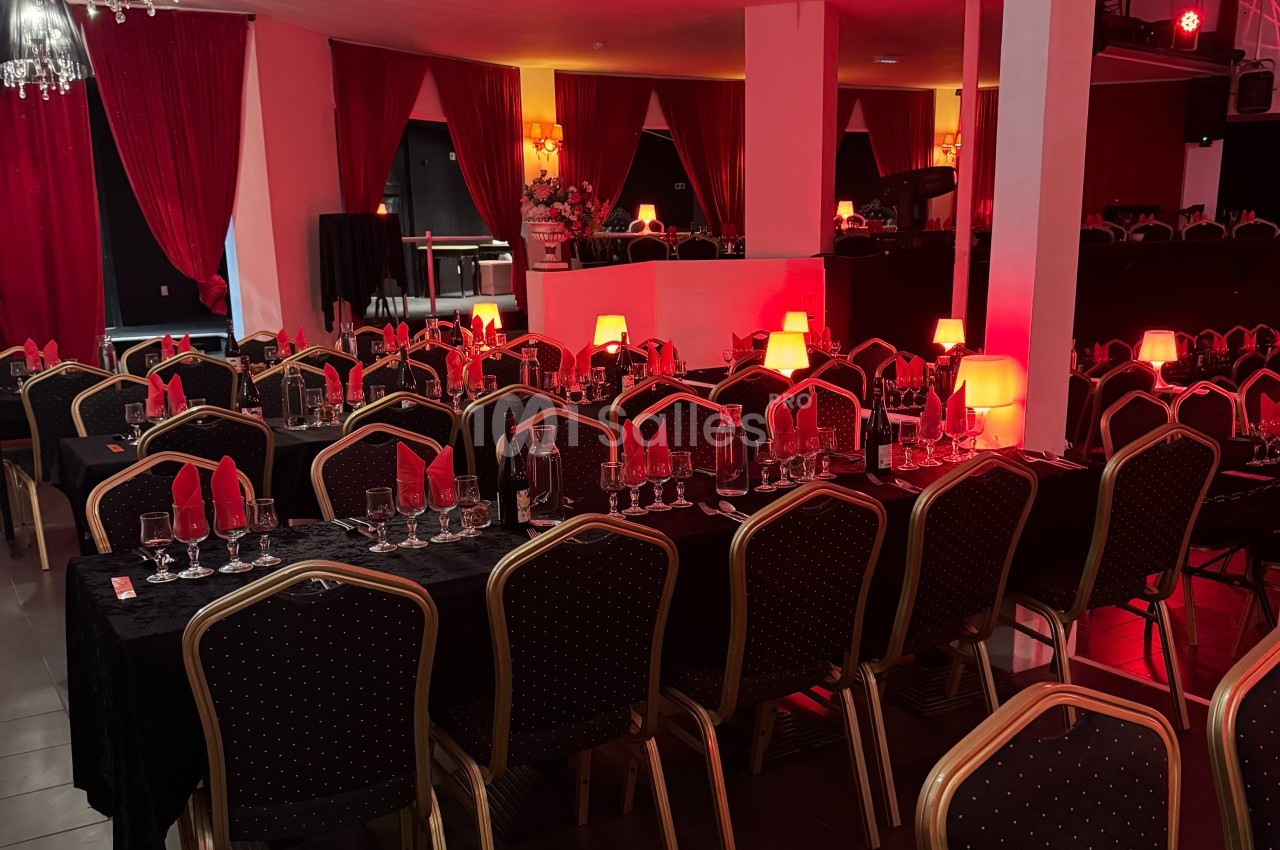 Salle de réception avec tables dressées, nappes noires, serviettes rouges et lampes diffusant une lumière tamisée.