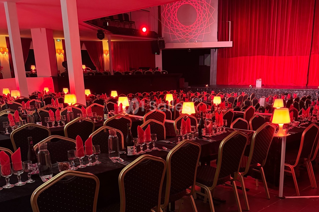 Salle de spectacle avec tables dressées, nappes noires, serviettes rouges et lampes diffusant une lumière tamisée.