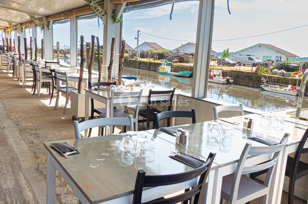 Terrasse d'un restaurant avec des tables dressées, donnant sur un port avec des bateaux et des cabanes en arrière-plan.
