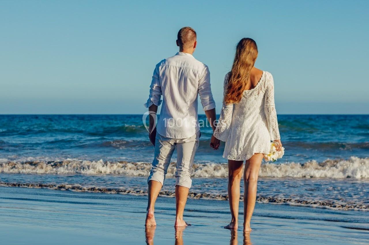 Un couple de dos, debout sur une plage, tenant la main, face à la mer sous un ciel dégagé.