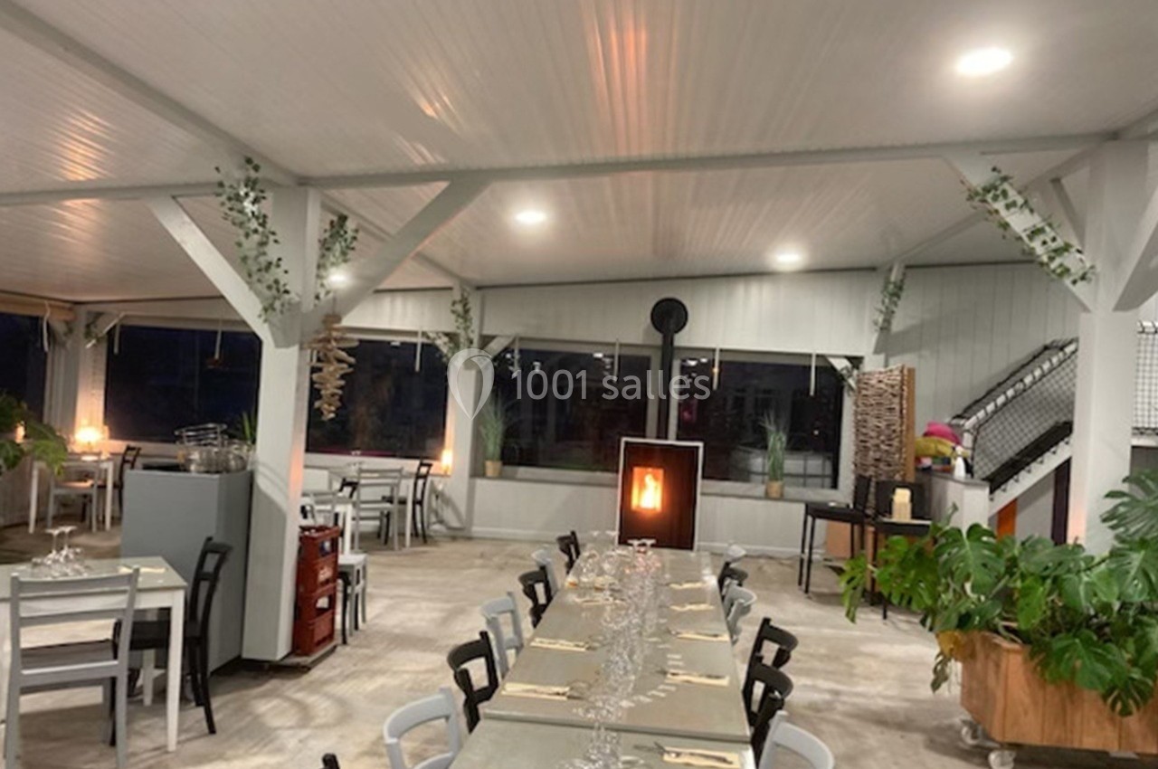 Salle de restaurant lumineuse avec une grande table dressée, poêle à bois allumé et décoration végétale.