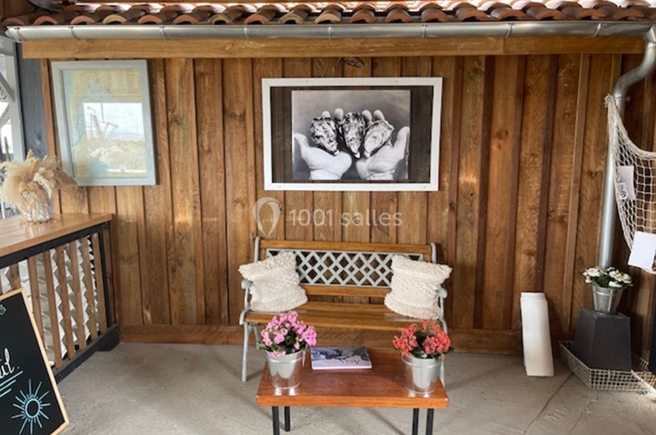 Banc en bois avec coussins, table basse avec pots de fleurs, et cadre photo sur un mur en bois dans un espace couvert.