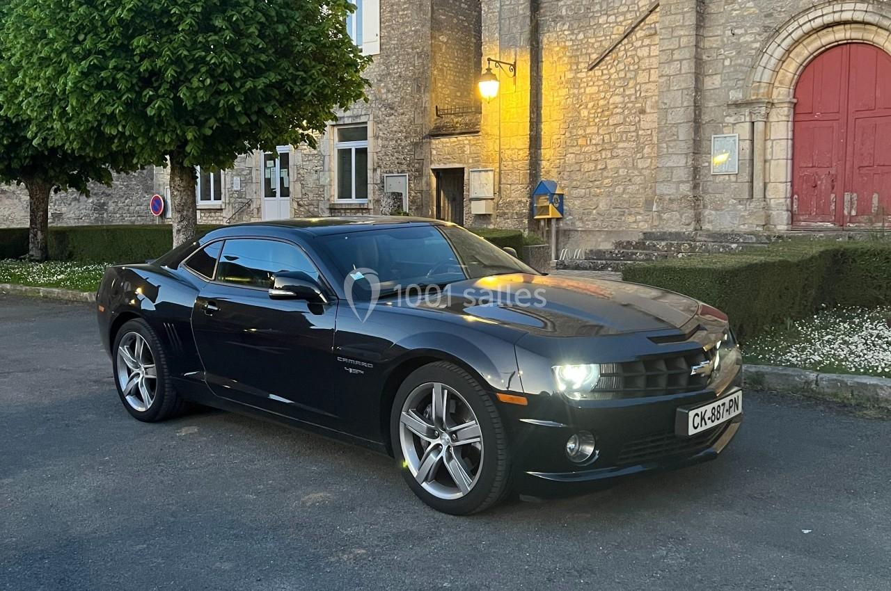 Voiture sportive noire garée devant un bâtiment en pierre avec une porte rouge et un éclairage de rue au crépuscule.