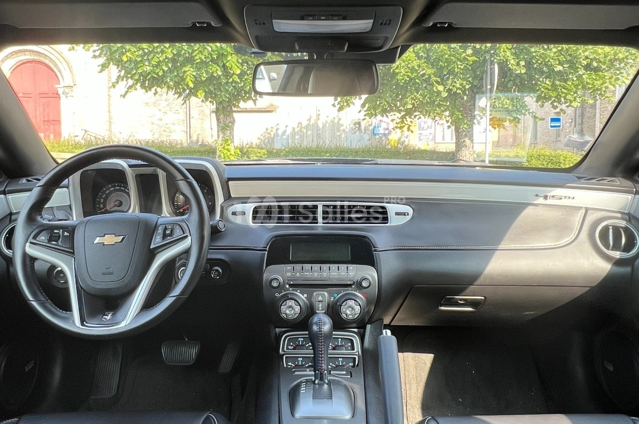 Vue de l'intérieur d'une voiture Chevrolet avec volant, tableau de bord et console centrale, devant un paysage urbain.