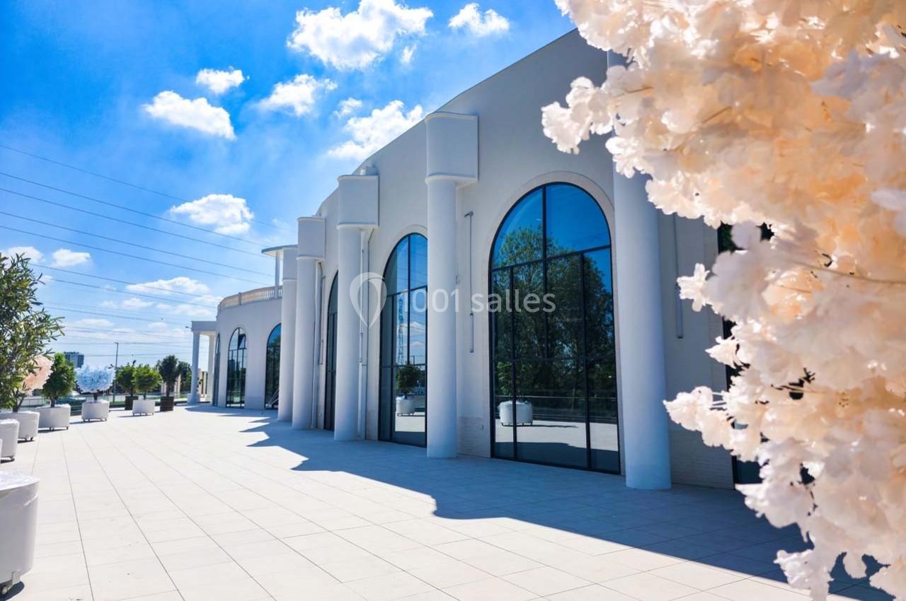 Façade d'un bâtiment moderne avec de grandes arches vitrées, entouré d'une terrasse ensoleillée et d'arbres décoratifs.