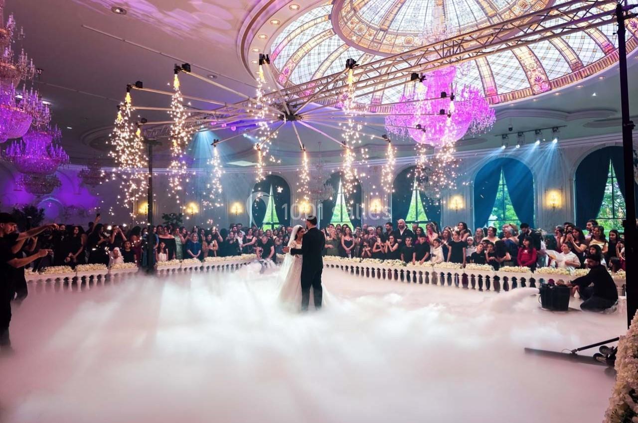 Un couple danse au centre d'une salle luxueuse avec un plafond en verre, éclairages scintillants et une foule en arrière…