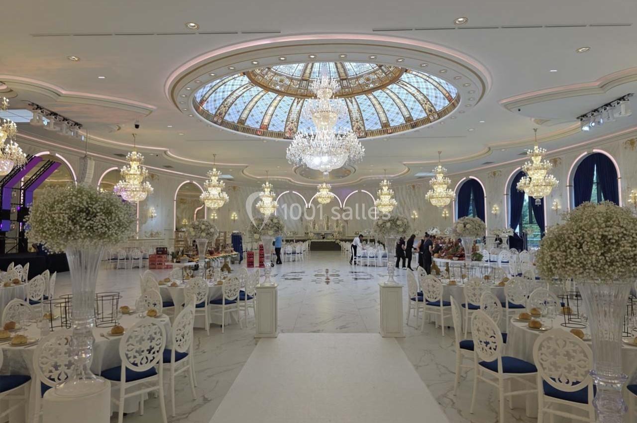 Salle de réception luxueuse avec lustres, plafond vitré, tables décorées et chaises blanches élégantes.