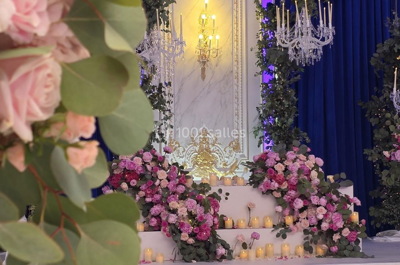 Décor floral avec roses roses et bougies sur des marches blanches, entouré de chandeliers et de rideaux bleus.
