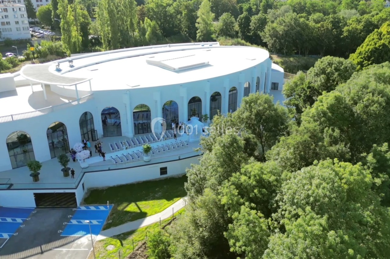 Vue aérienne d'un bâtiment blanc moderne entouré de verdure, avec une terrasse aménagée et des personnes présentes.