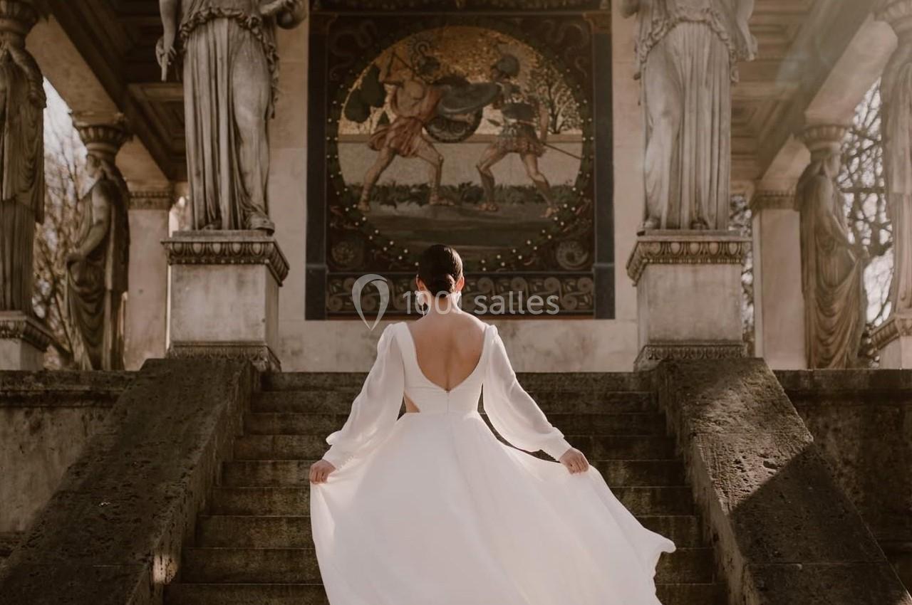Femme en robe blanche montant un escalier en pierre, devant une structure ornée de statues et d'une mosaïque.