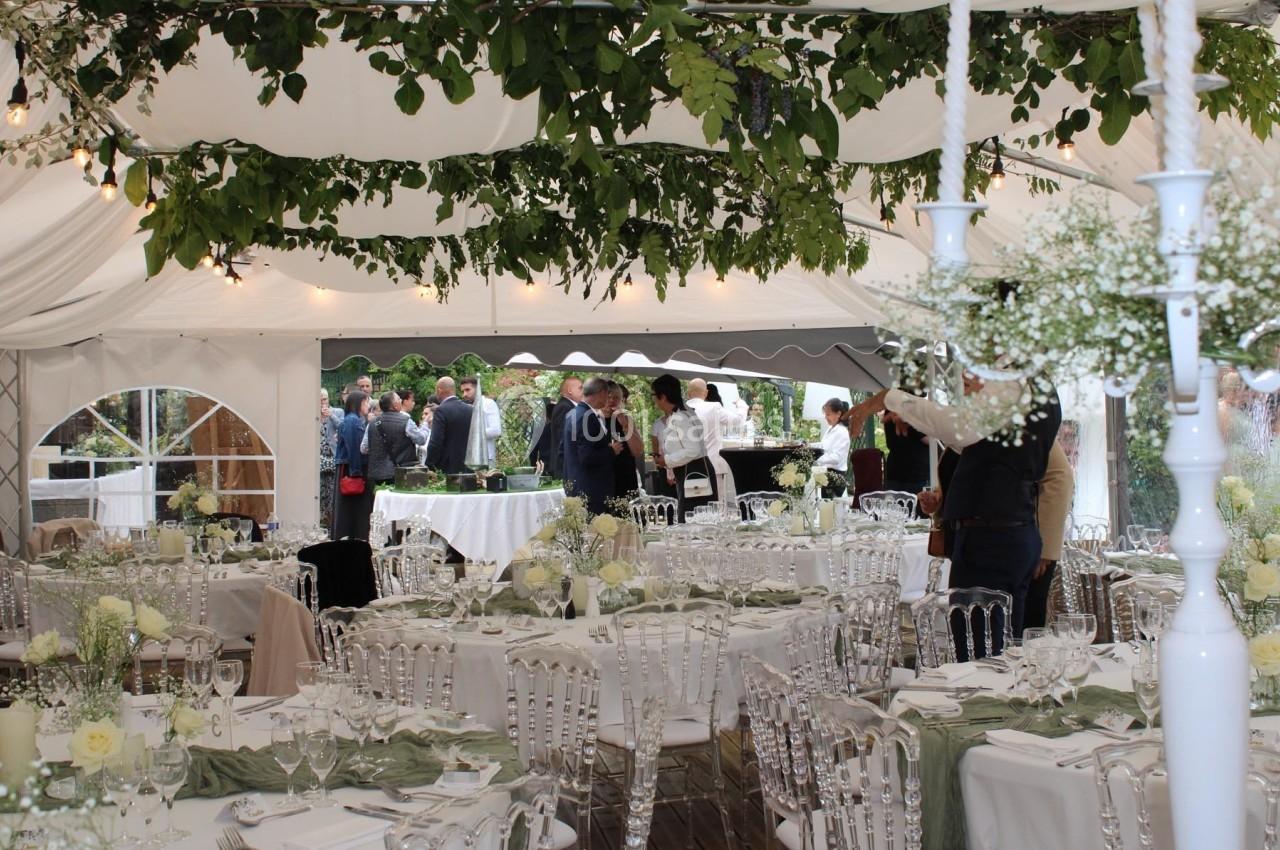 Salle de réception sous une tente décorée de feuillage, avec tables dressées et invités en arrière-plan.