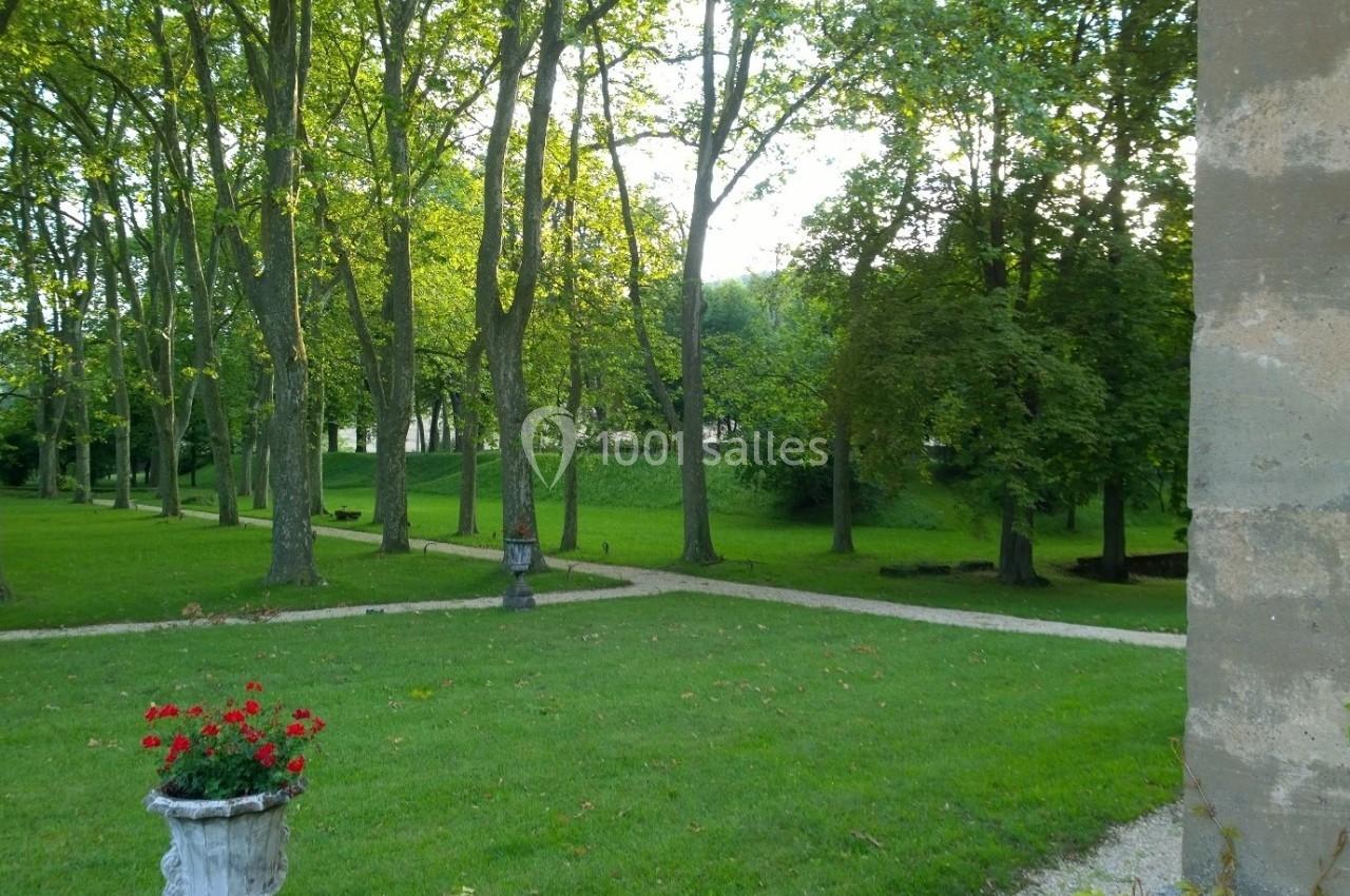 Allée bordée d'arbres dans un parc verdoyant, avec un pot de fleurs rouges au premier plan.