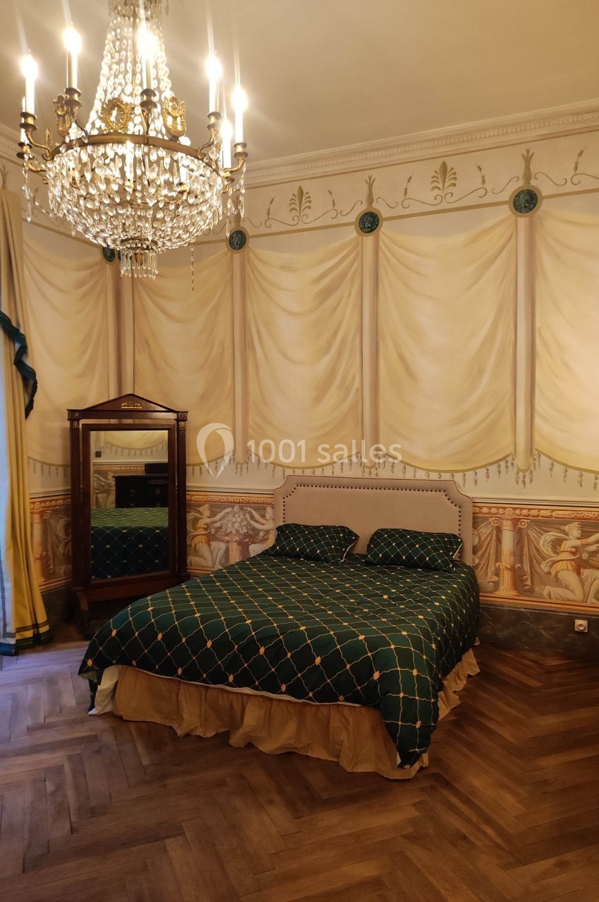 Chambre avec lit double, draperies murales peintes, parquet en bois, armoire ancienne et lustre en cristal.