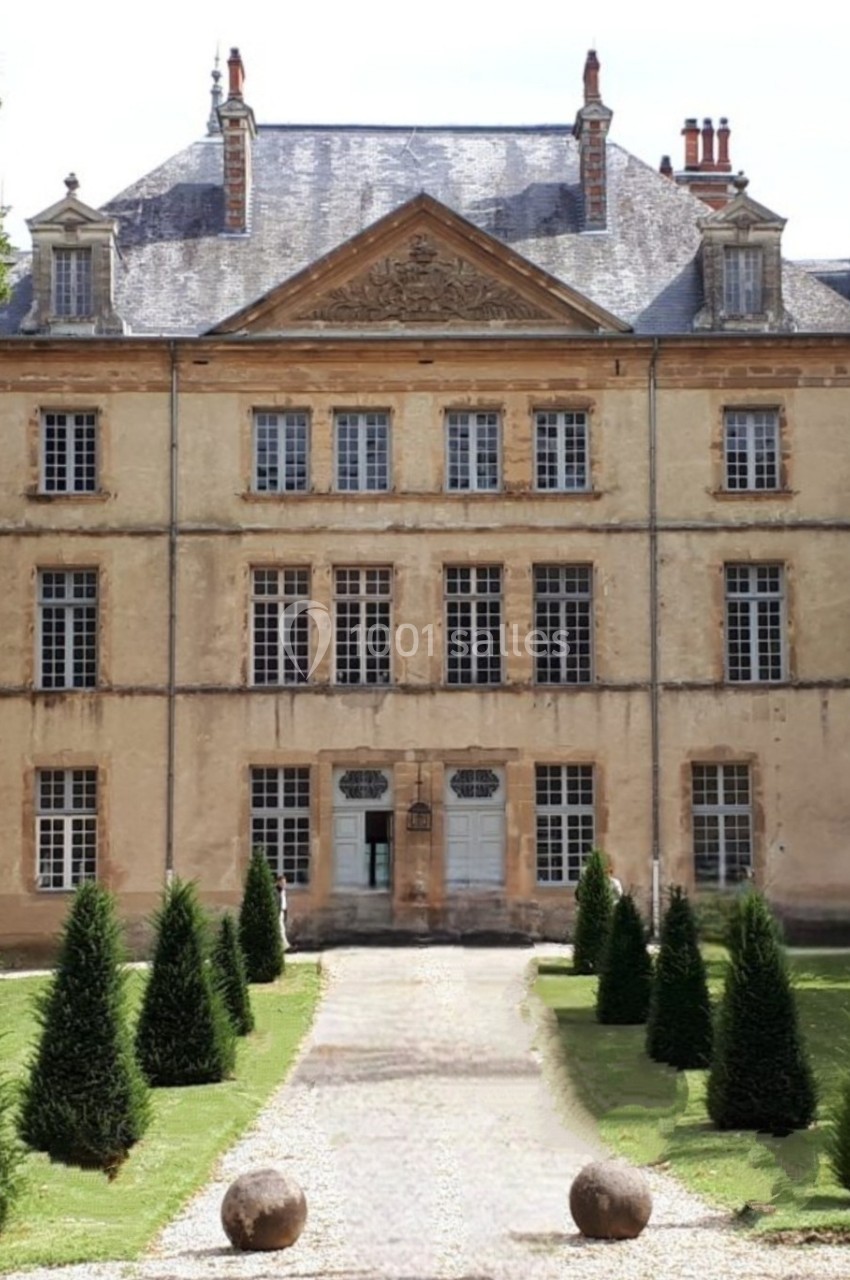 Façade d'un manoir ancien en pierre, entouré d'un jardin avec allée centrale bordée d'arbustes taillés.