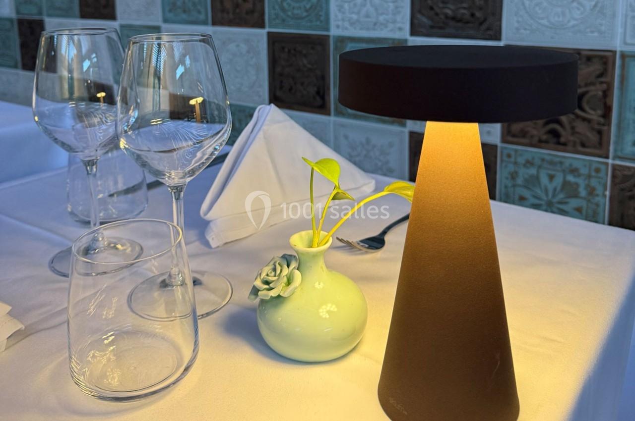Table dressée avec des verres, une serviette pliée, une petite lampe et un vase contenant une plante.