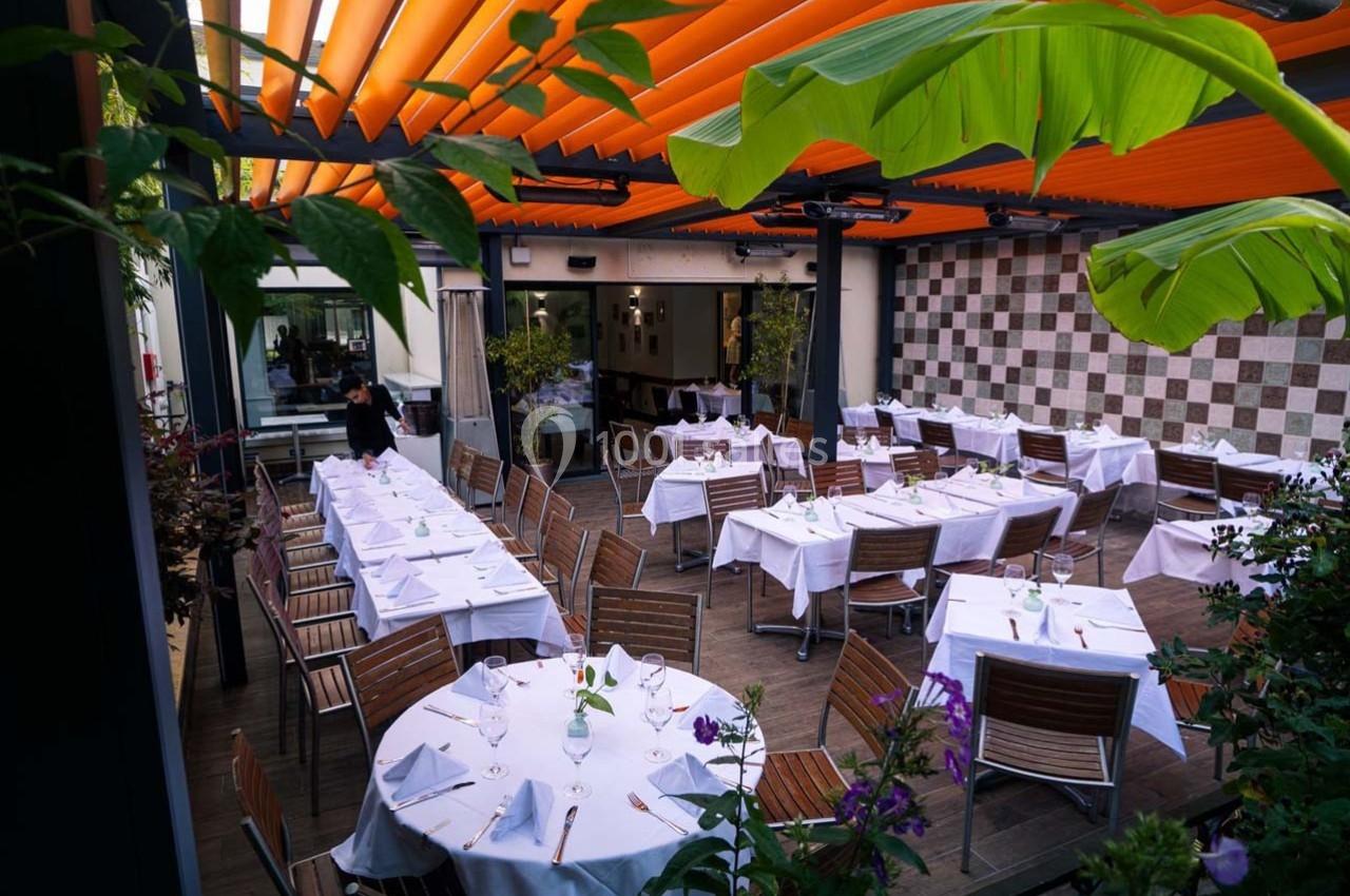 Terrasse de restaurant avec tables dressées, chaises en bois, pergola orange et plantes décoratives.