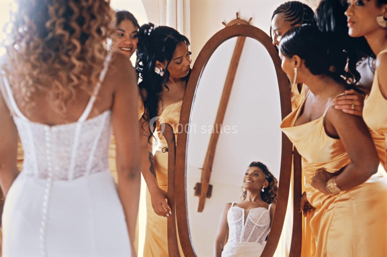 Une mariée en robe blanche se regarde dans un miroir entourée de demoiselles d'honneur en robes jaunes.