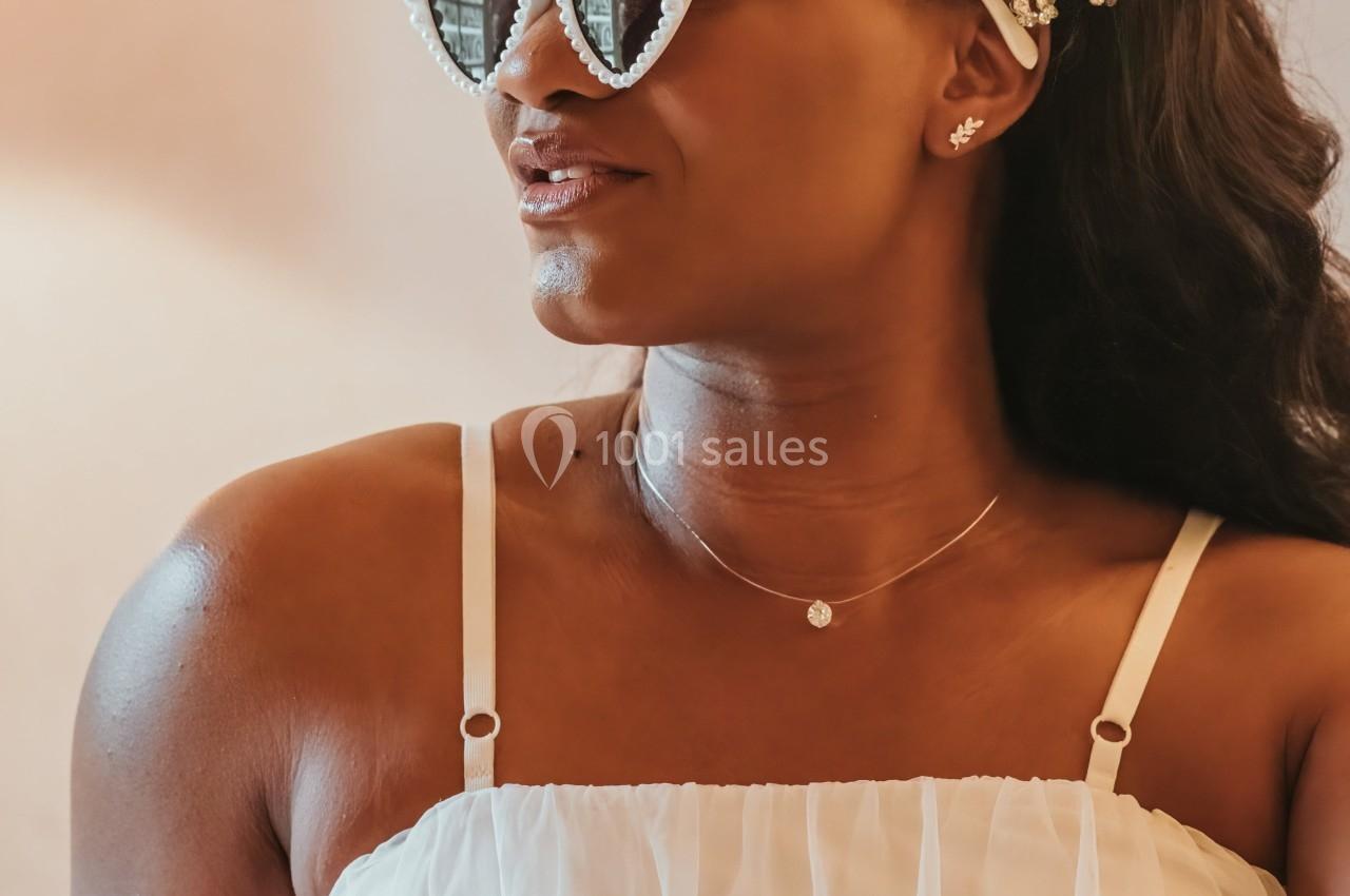 Femme portant une robe blanche, des lunettes en forme de cœur et un serre-tête orné de perles, assise dans un intérieur…