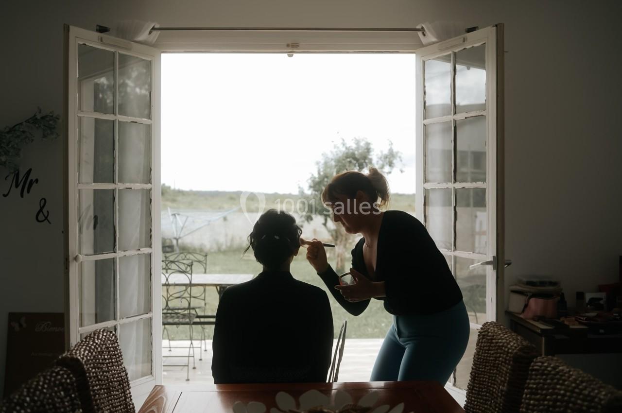 Une maquilleuse applique du maquillage sur une personne assise près d'une fenêtre ouverte donnant sur un jardin.