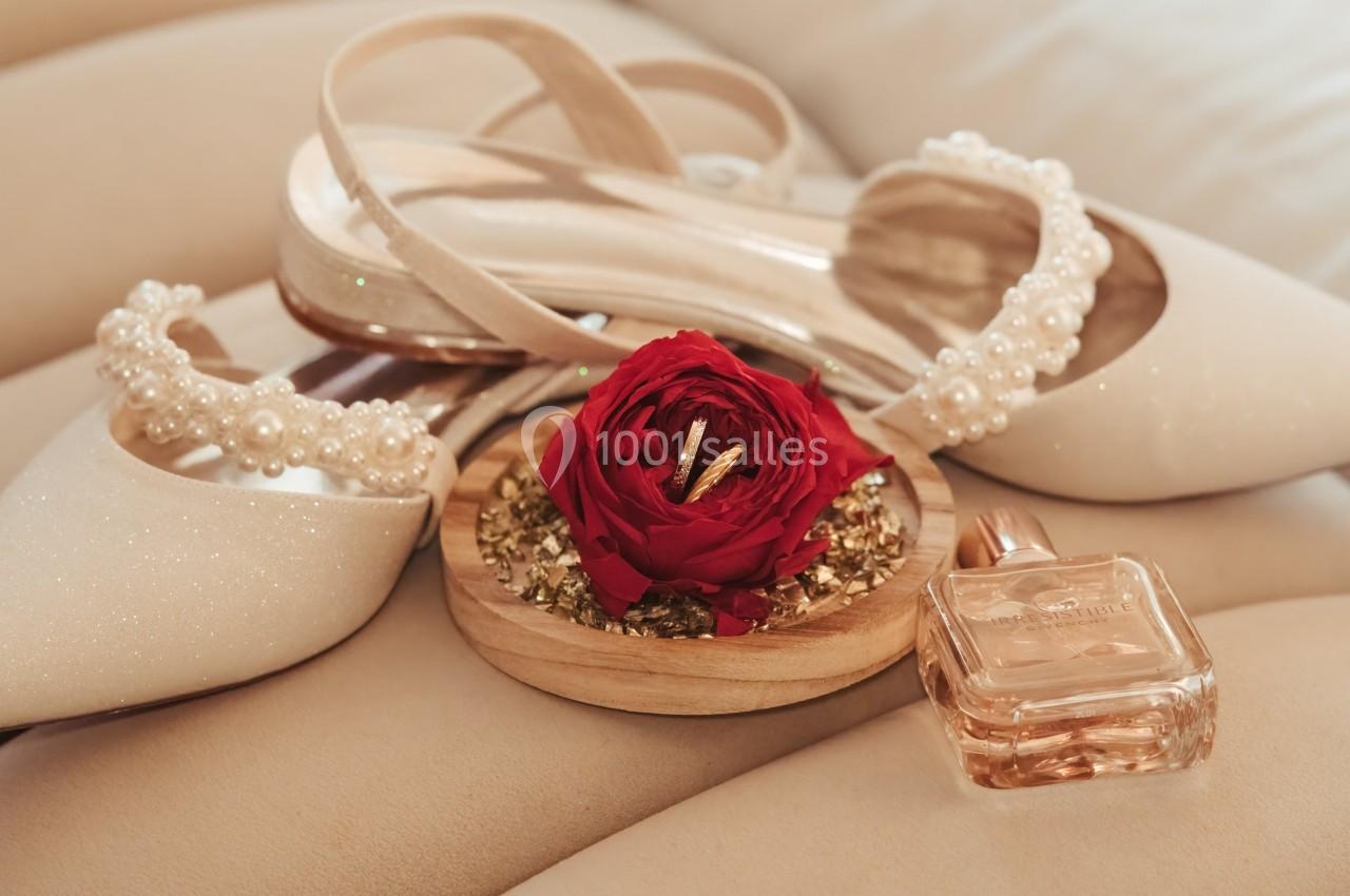 Chaussures blanches ornées de perles, rose rouge et flacons de parfum sur un coussin beige.