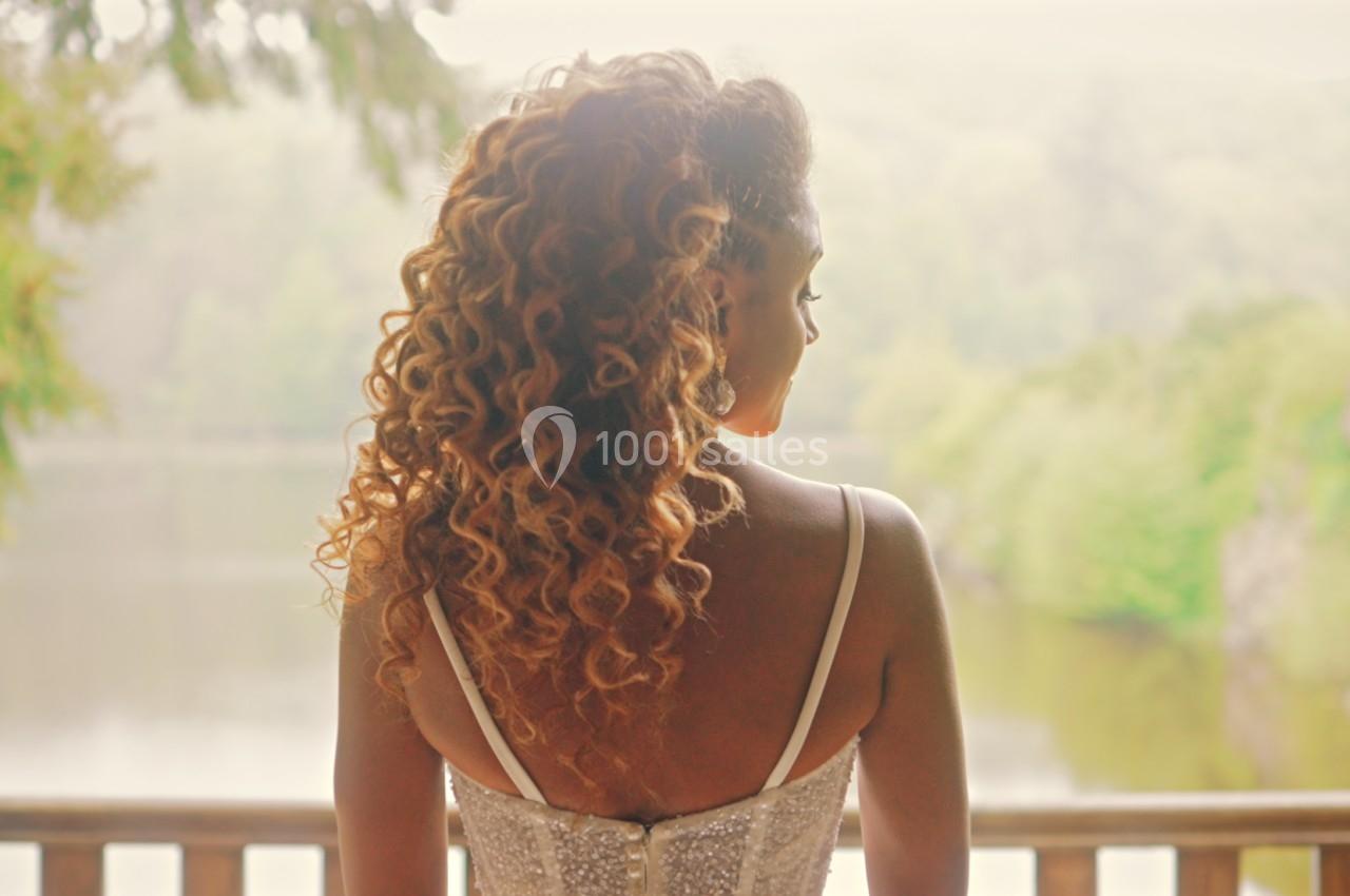 Femme de dos avec des cheveux bouclés, regardant un lac entouré de verdure depuis un balcon en bois.